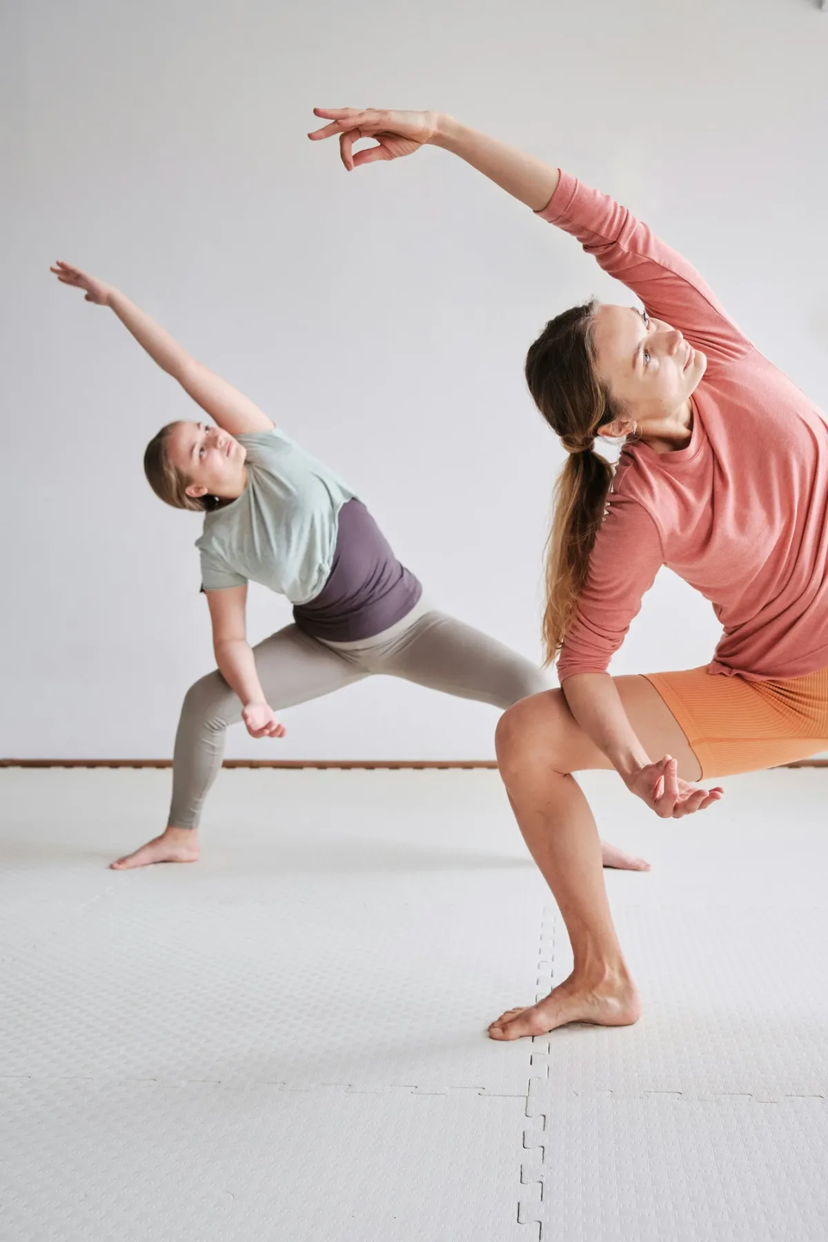 Zwei Frauen praktizieren gemeinsam Yoga in einer Seitendehnung auf wei&szlig;em Boden.