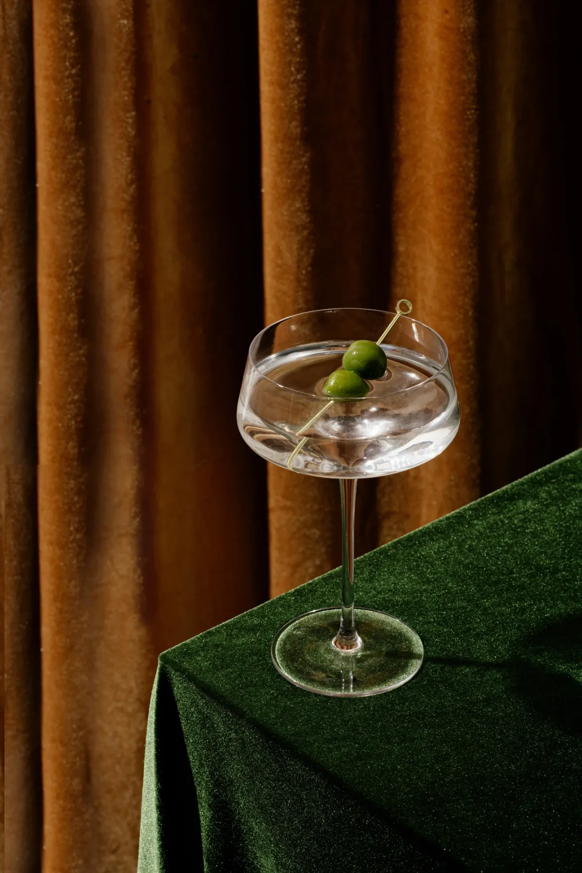Elegantes Cocktailglas mit Eis und gr&uuml;ner Olive auf gr&uuml;nem Samt-Tischtuch vor goldenem Vorhang.
