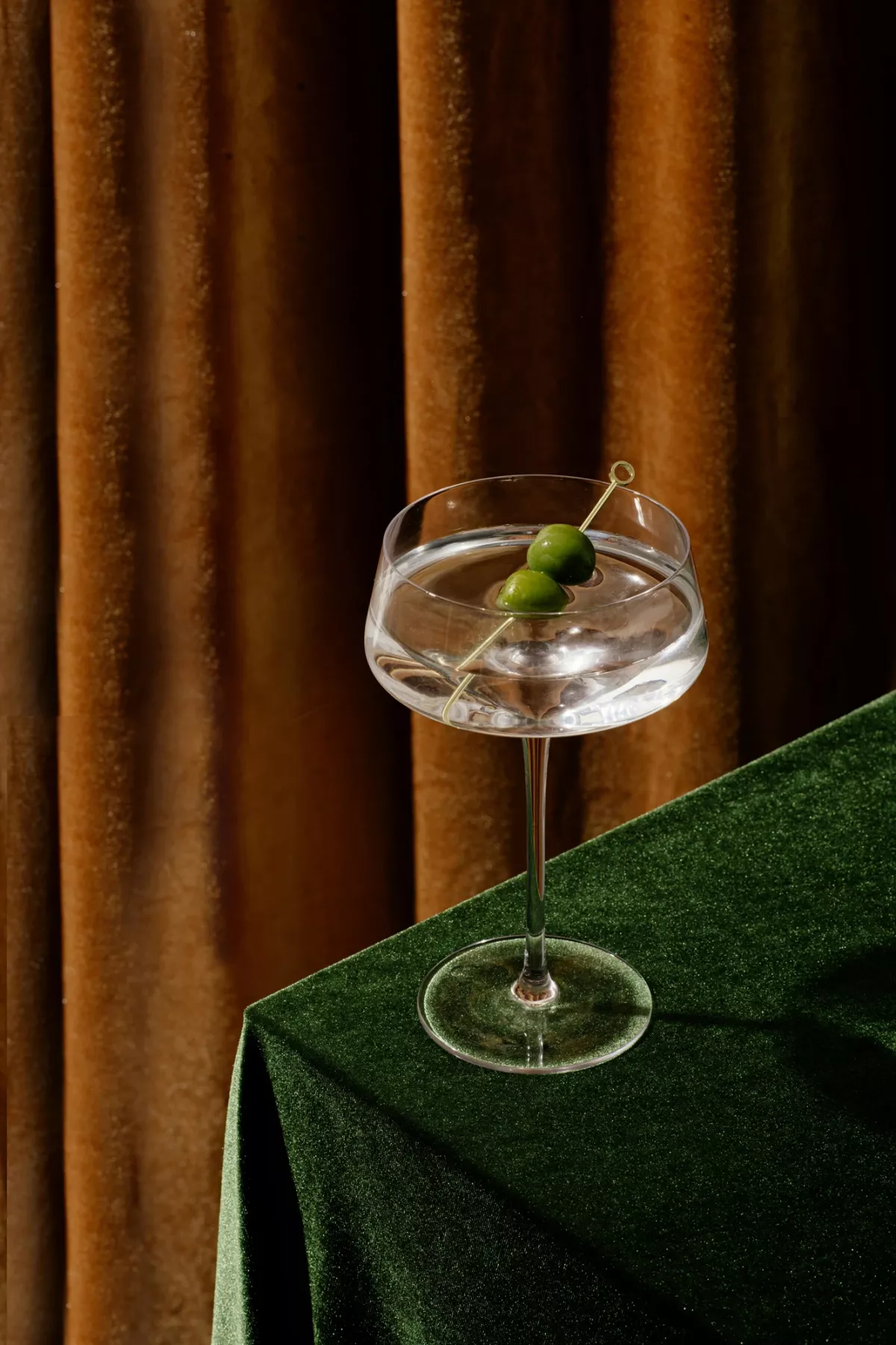 Elegantes Cocktailglas mit Eis und gr&uuml;ner Olive auf gr&uuml;nem Samt-Tischtuch vor goldenem Vorhang.