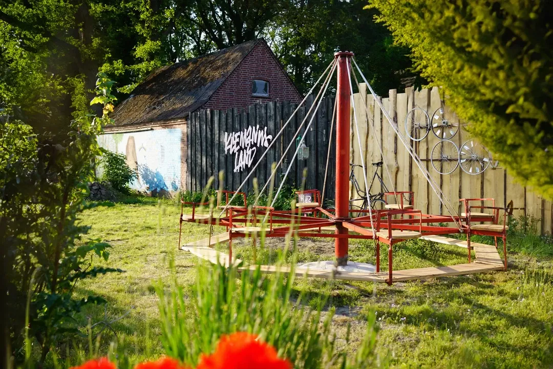 Rustikale Scheune mit Schriftzug und roten Metallkonstruktionen auf gr&uuml;ner Wiese, umgeben von Hecken und B&auml;umen.