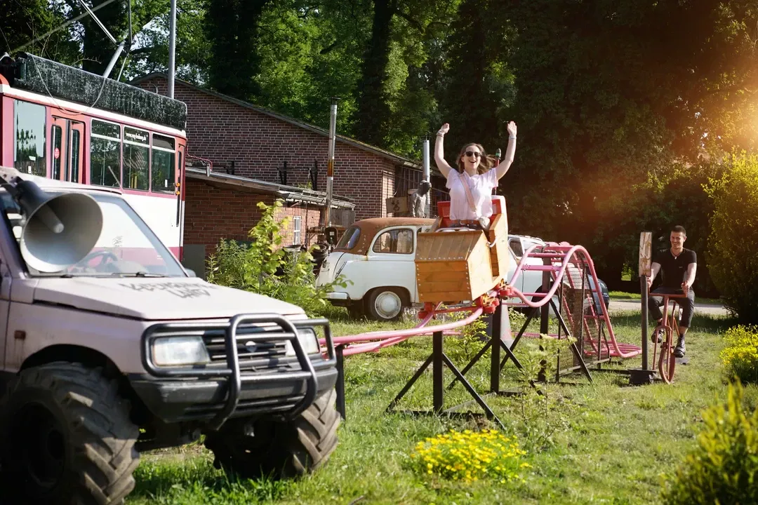 Teambuilding-Aktivit&auml;t auf dem Gel&auml;nde: Person jubelt auf einer improvisierten Achterbahn, w&auml;hrend ein Gel&auml;ndewagen und Fahrr&auml;der in Umgebung bereitstehen.