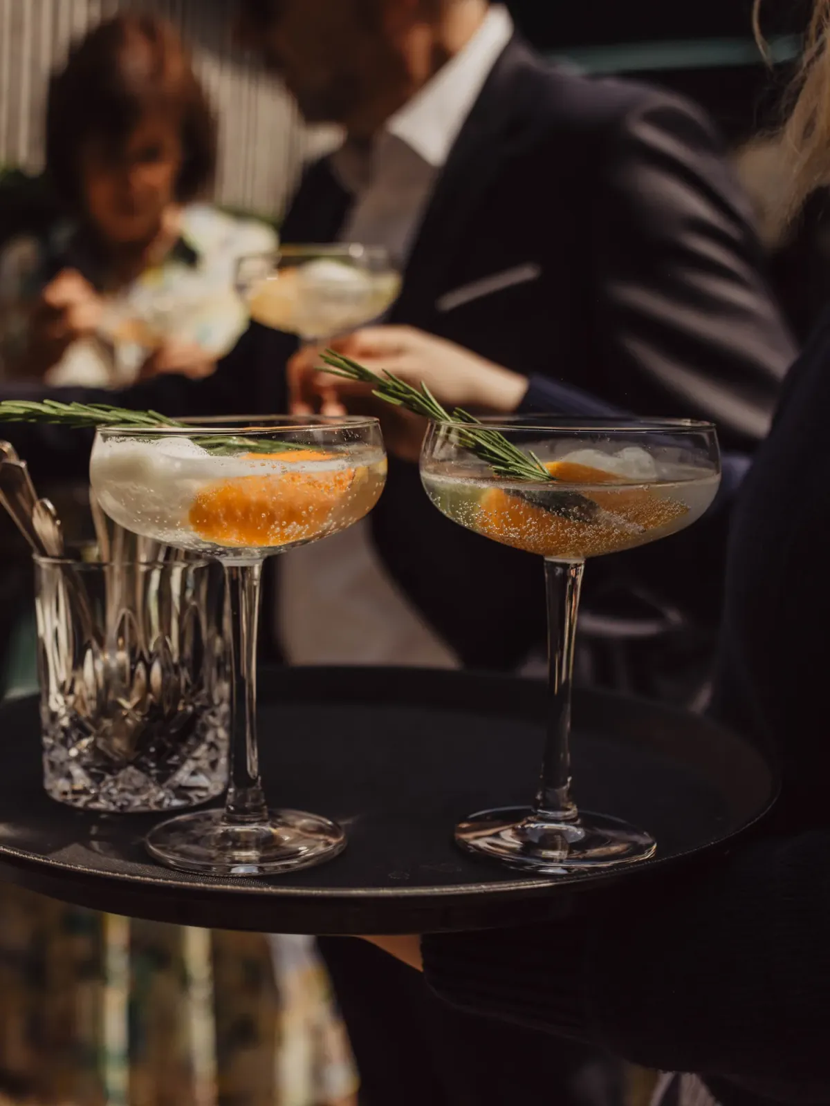 Zwei elegante Cocktails mit Rosmarinkr&auml;utern auf einem schwarzen Tablett bei einer Feier, im Hintergrund G&auml;ste in formaler Kleidung.