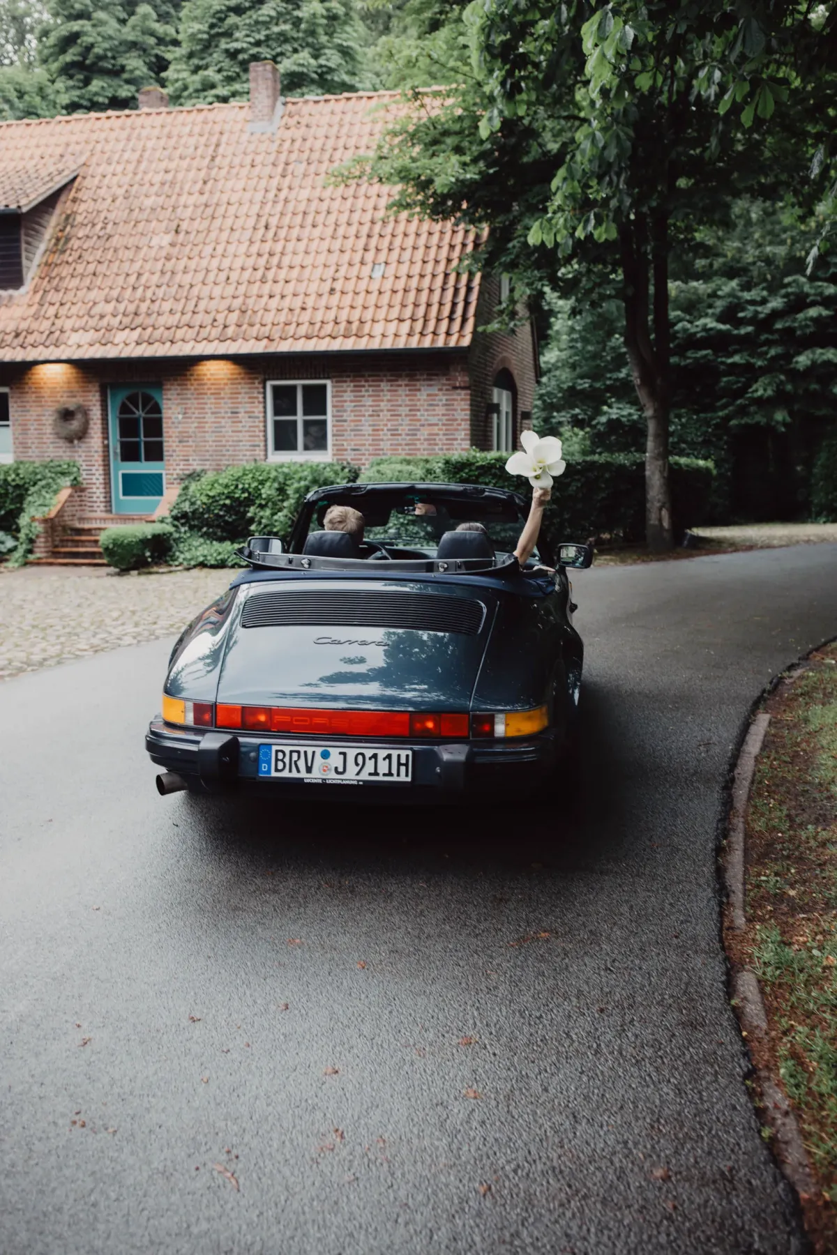 Schwarzer Porsche 911 Cabriolet mit Dachgep&auml;ck f&auml;hrt vor Backsteinfachwerk-Haus umgeben von gr&uuml;nem Garten.