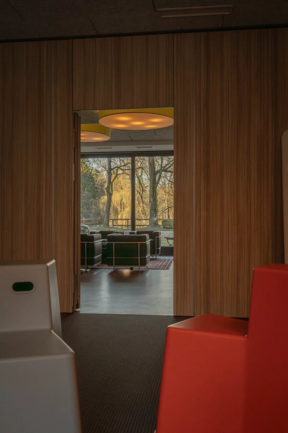 Blick durch offene T&uuml;r aus modernen Veranstaltungsraum mit roten und wei&szlig;en Sitzelementen, im Hintergrund Ledersessel vor gro&szlig;er Fensterscheibe mit Blick auf B&auml;ume und Teich.