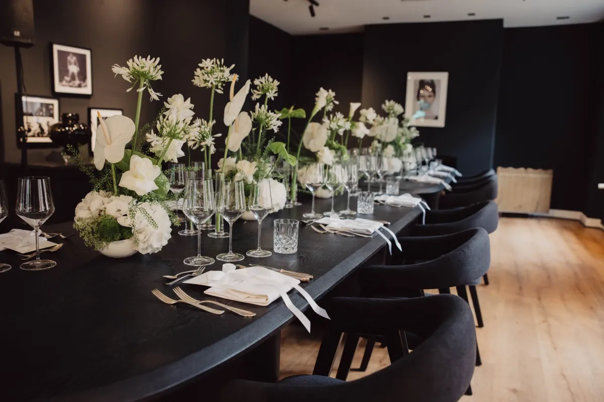 Elegant gedeckter Tisch mit schwarzer Tischplatte, wei&szlig;en Blumenarrangements in Glasvasen und schwarzen St&uuml;hlen in minimalistischem Veranstaltungsraum.