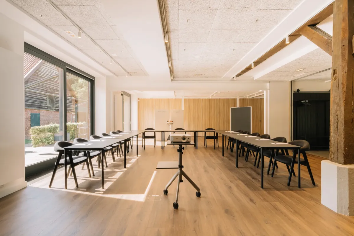Heller Veranstaltungsraum mit Holzfu&szlig;boden, modernen schwarzen St&uuml;hlen, gro&szlig;en Fenstern und Holzbalken an der Decke, ideal f&uuml;r Konferenzen und Firmenfeiern.