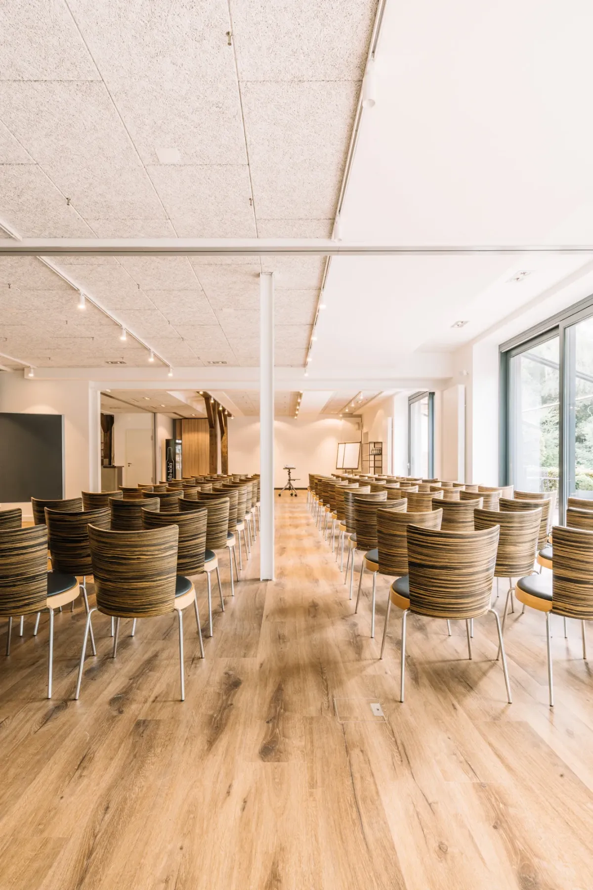 Moderner Veranstaltungsraum mit Holzfu&szlig;boden, Reihen von modernen St&uuml;hlen mit Holzschalen und gro&szlig;en Fenstern, die nat&uuml;rliches Licht spenden.