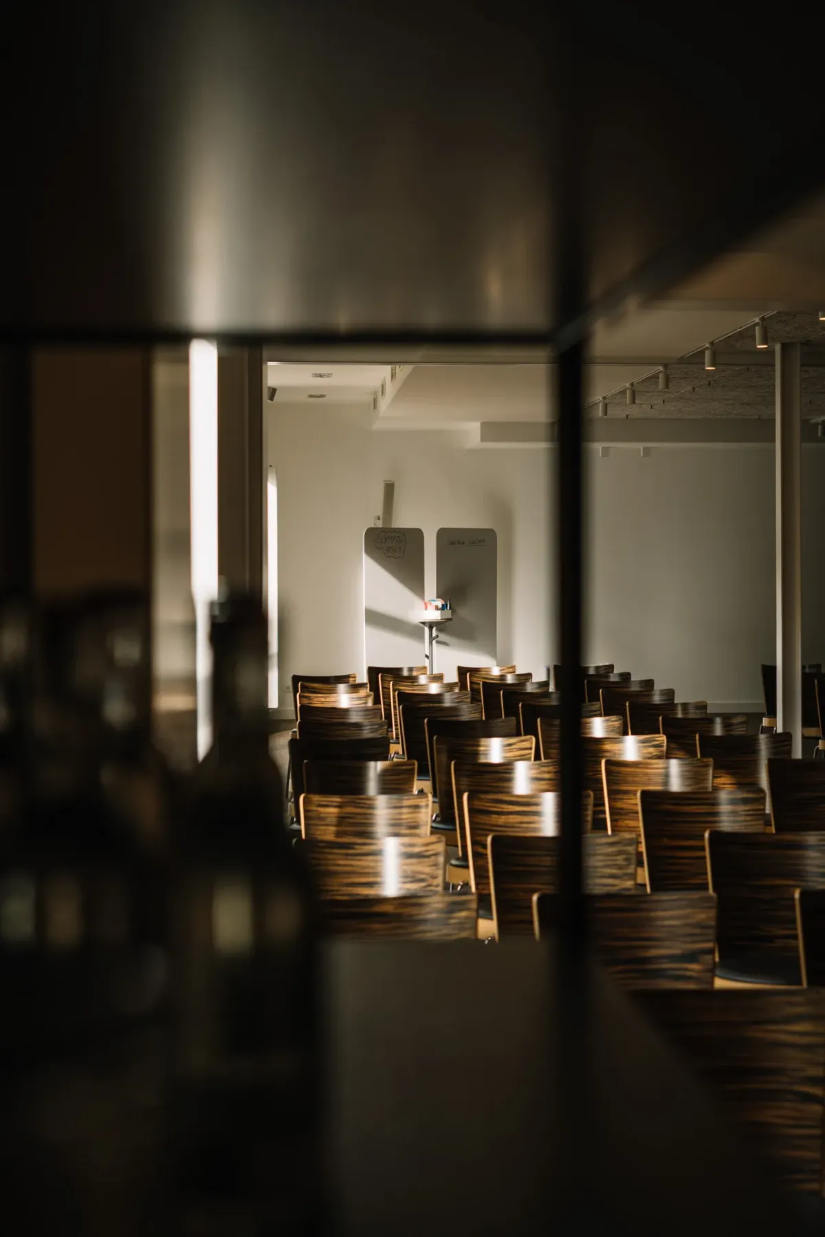Blick durch offene Glast&uuml;ren in einen modernen Seminarraum mit Holzst&uuml;hlen, warm beleuchtet durch Licht von drau&szlig;en.