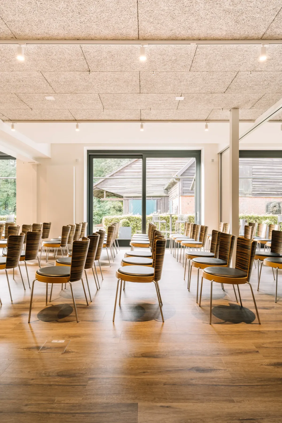 Moderner Veranstaltungsraum mit Reihen von Holzst&uuml;hlen, gro&szlig;en Fenstern mit Blick ins Gr&uuml;ne und heller Decke, ideal f&uuml;r Hochzeiten und Firmenevents.