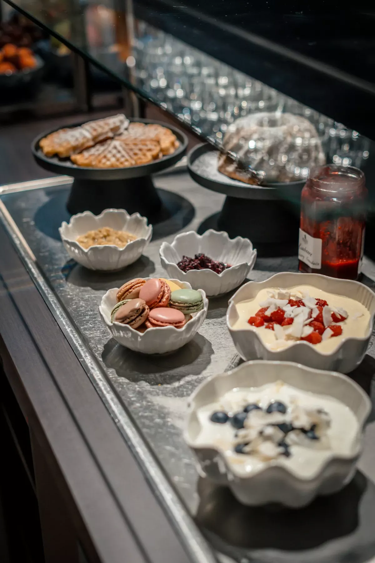Fr&uuml;hst&uuml;ckstheke mit verschiedenen Toppings in wei&szlig;en Schalen: N&uuml;sse, Fr&uuml;chte, Schokolade und Sahne, im Hintergrund Waffeleisen.