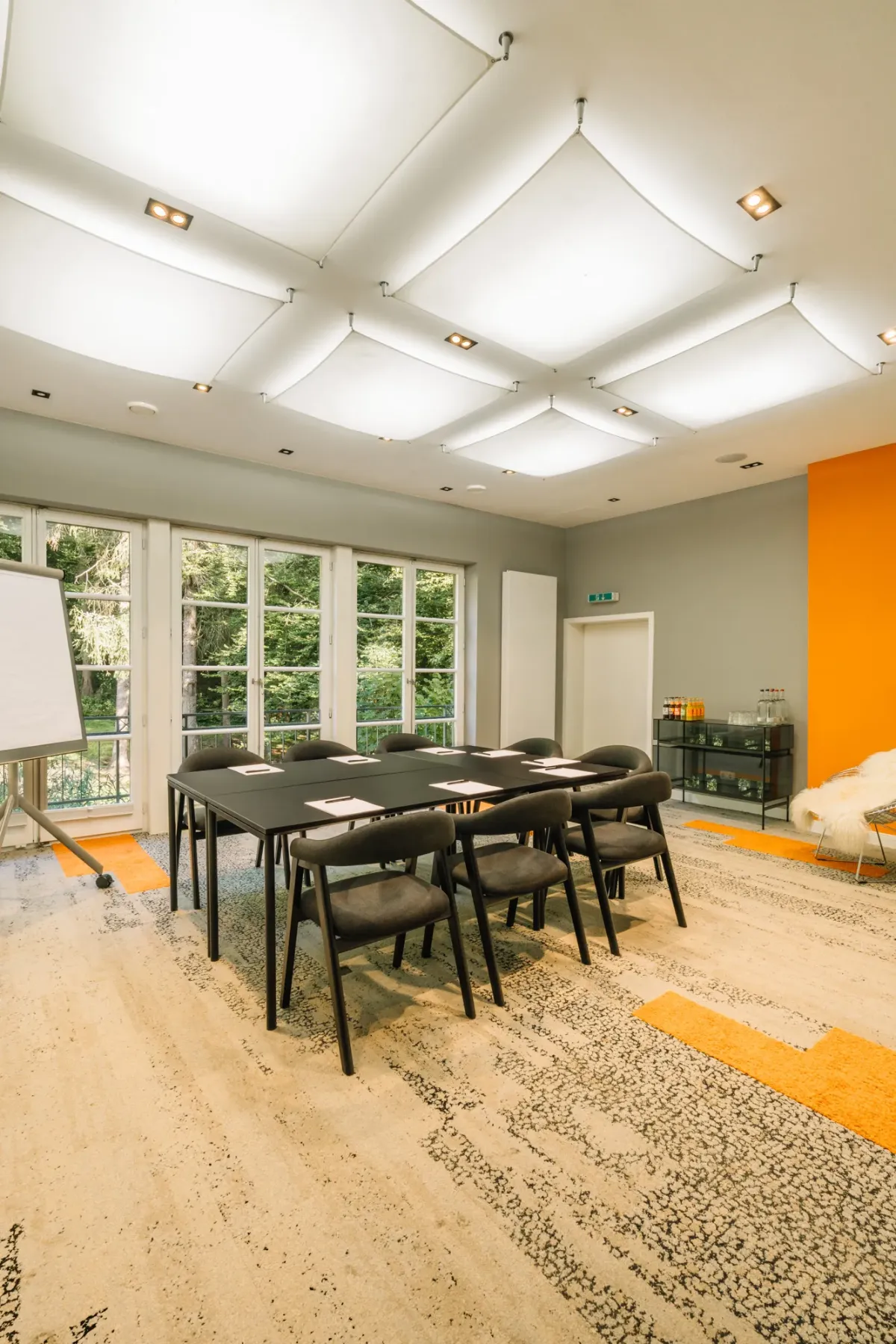 Moderner Seminarraum mit gro&szlig;z&uuml;gigen Fenstern, schwarzem Konferenztisch, ergonomischen St&uuml;hlen, Flipchart und orange-grauen Designelementen.
