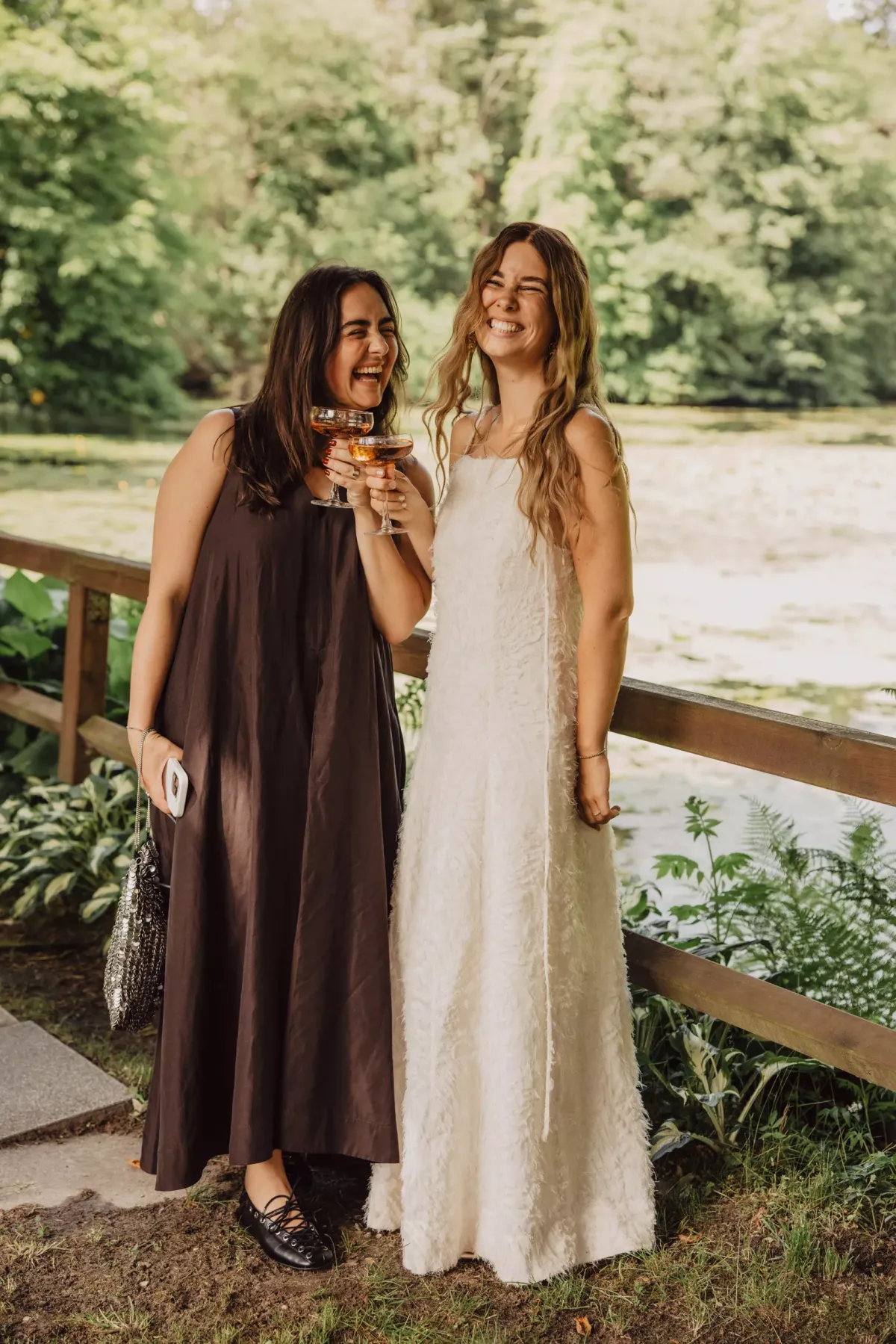 Zwei Frauen stehen an einem Holzgel&auml;nder vor gr&uuml;ner Naturkulisse. Die eine tr&auml;gt ein braunes, die andere ein cremefarbenes langes Kleid.