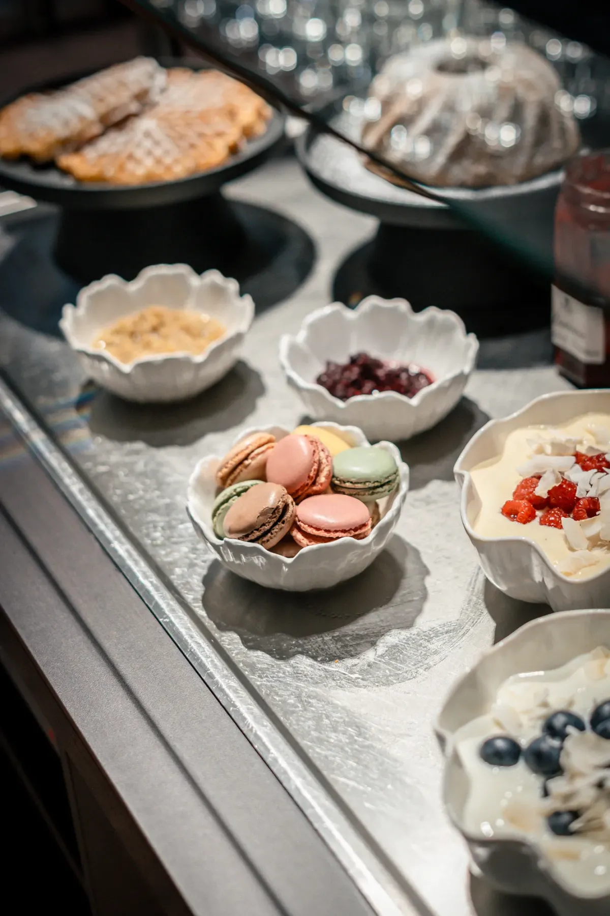 Elegante Fr&uuml;hst&uuml;cks-Buffet mit wei&szlig;en Sch&auml;lchen: Pistazien, rote Beeren, N&uuml;sse und Fr&uuml;chte neben Geb&auml;ck und Kuchen auf silberner Fl&auml;che.