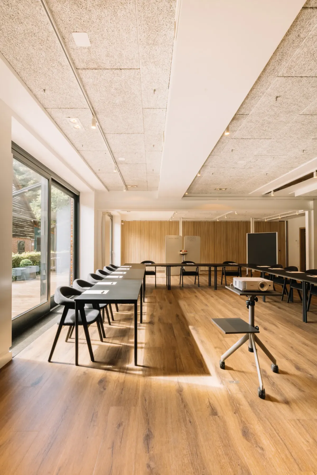 Moderner Veranstaltungsraum mit Holzparkett, hellen Decken, gro&szlig;en Fenstern und flexibel arrangierten schwarzen St&uuml;hlen sowie Tischen f&uuml;r Events und Meetings.