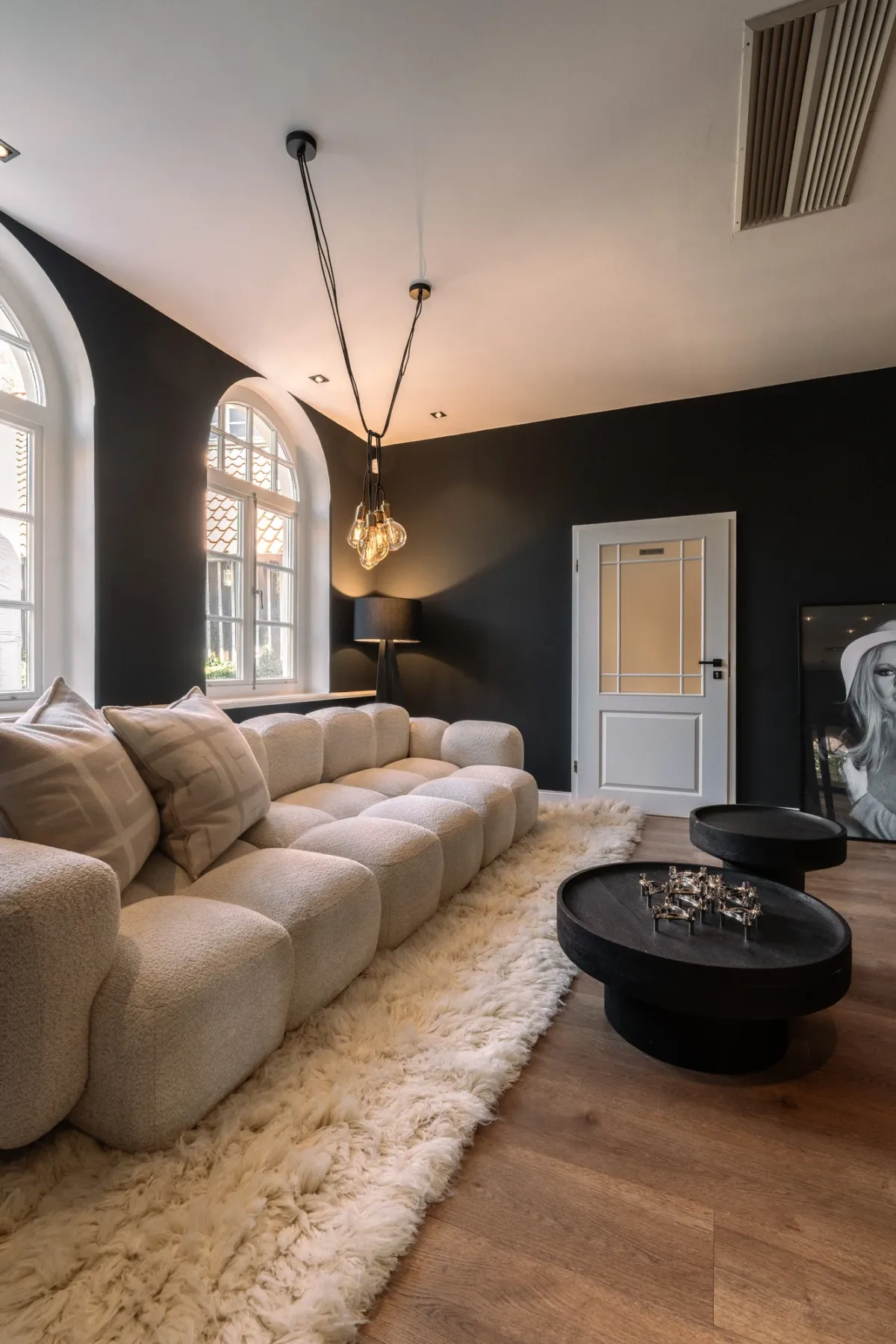 Modernes Zimmer mit cremewei&szlig;em Sofa, schwarzer Wand, gro&szlig;em Rundbogenfenster und eleganter Pendelleuchte &uuml;ber ovalem schwarzem Couchtisch.