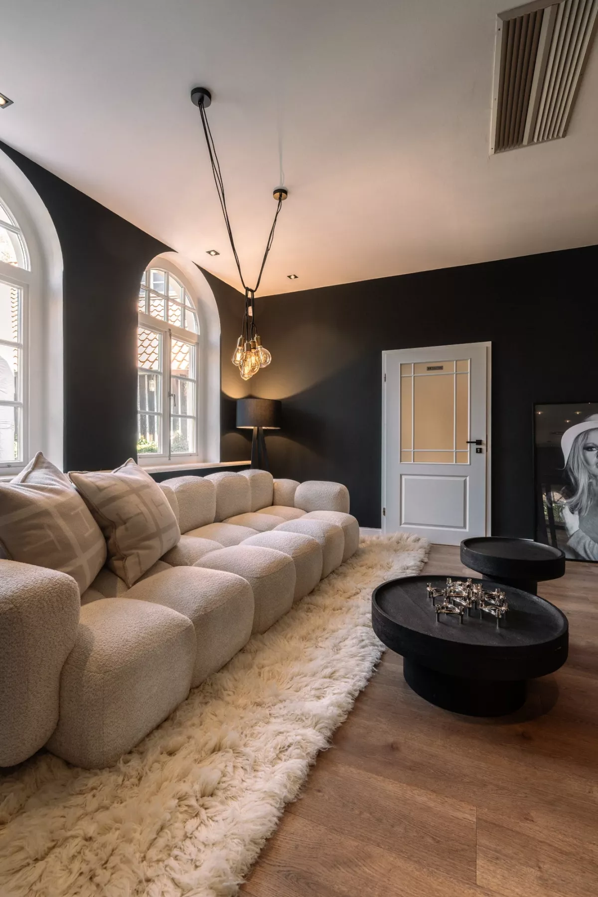 Modernes Zimmer mit cremewei&szlig;em Sofa, schwarzer Wand, gro&szlig;em Rundbogenfenster und eleganter Pendelleuchte &uuml;ber ovalem schwarzem Couchtisch.
