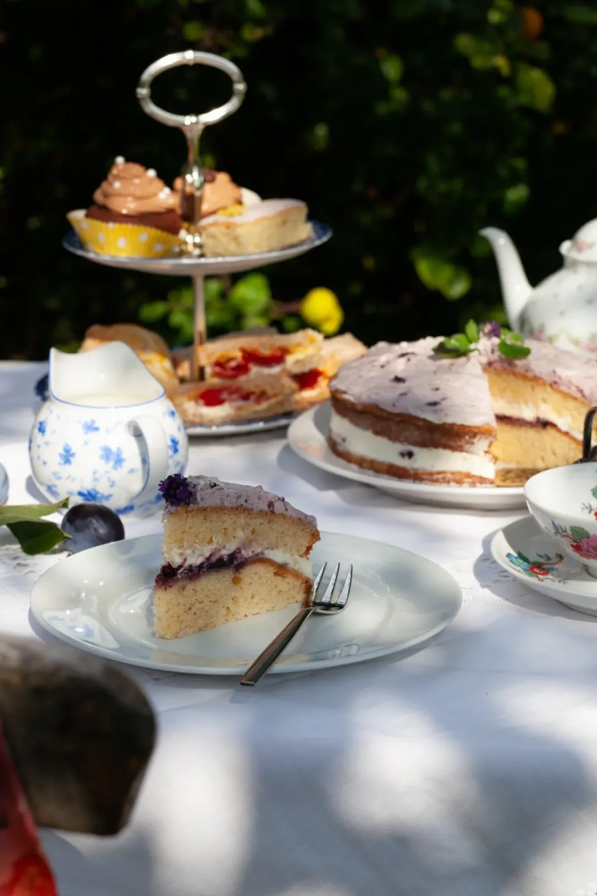 Elegante Kaffeetafel im Garten mit mehrschichtigem Kuchenst&auml;nder, Tortenscheiben auf Tellern und Teservice vor gr&uuml;nem Hintergrund.