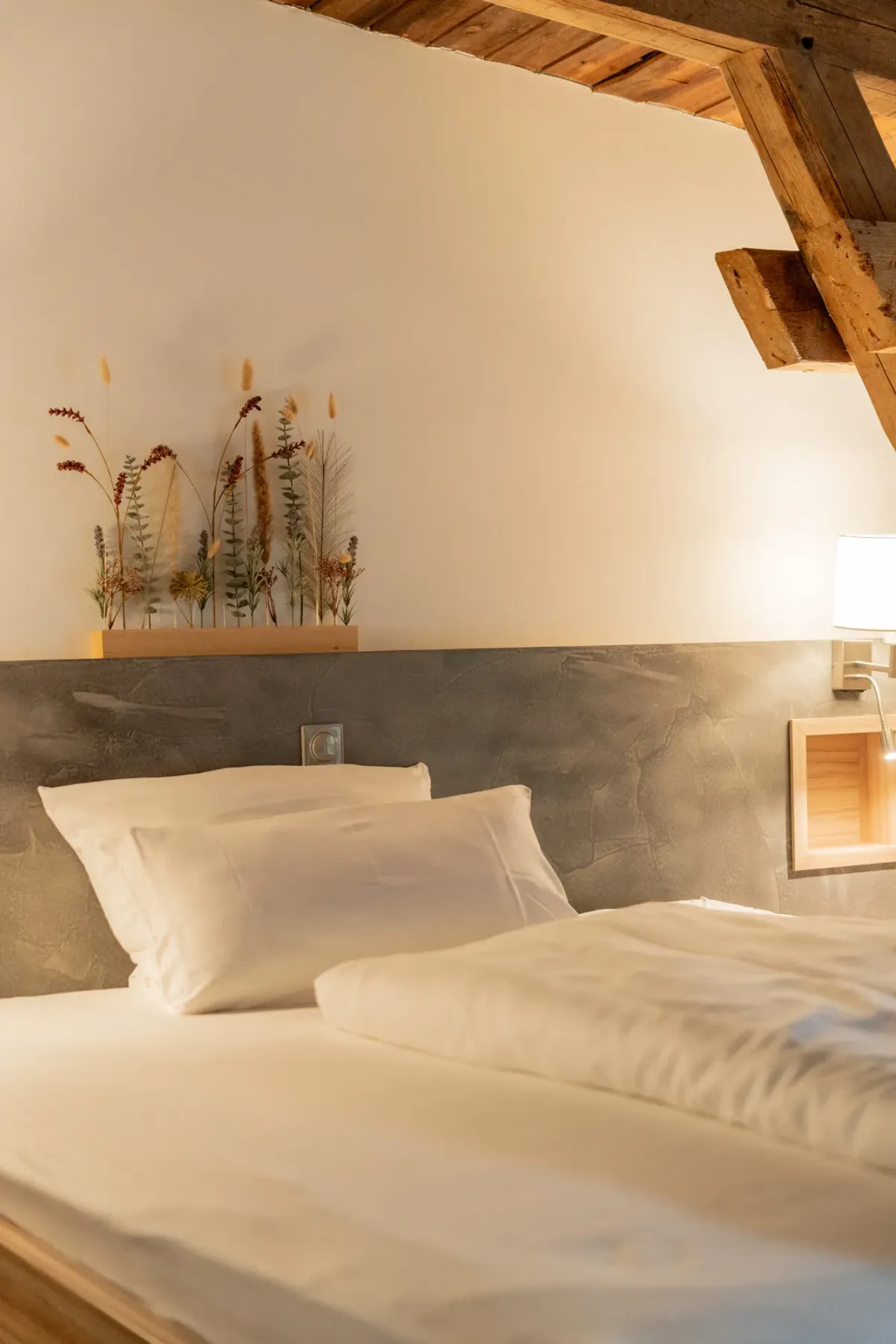 Gem&uuml;tliches Schlafzimmer mit wei&szlig;er Bettw&auml;sche, grauem Kopfteil und rustikalen Holzbalken an der Decke.