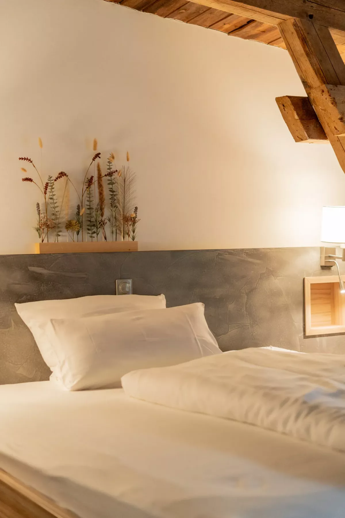 Gem&uuml;tliches Schlafzimmer mit wei&szlig;er Bettw&auml;sche, grauem Kopfteil und rustikalen Holzbalken an der Decke.