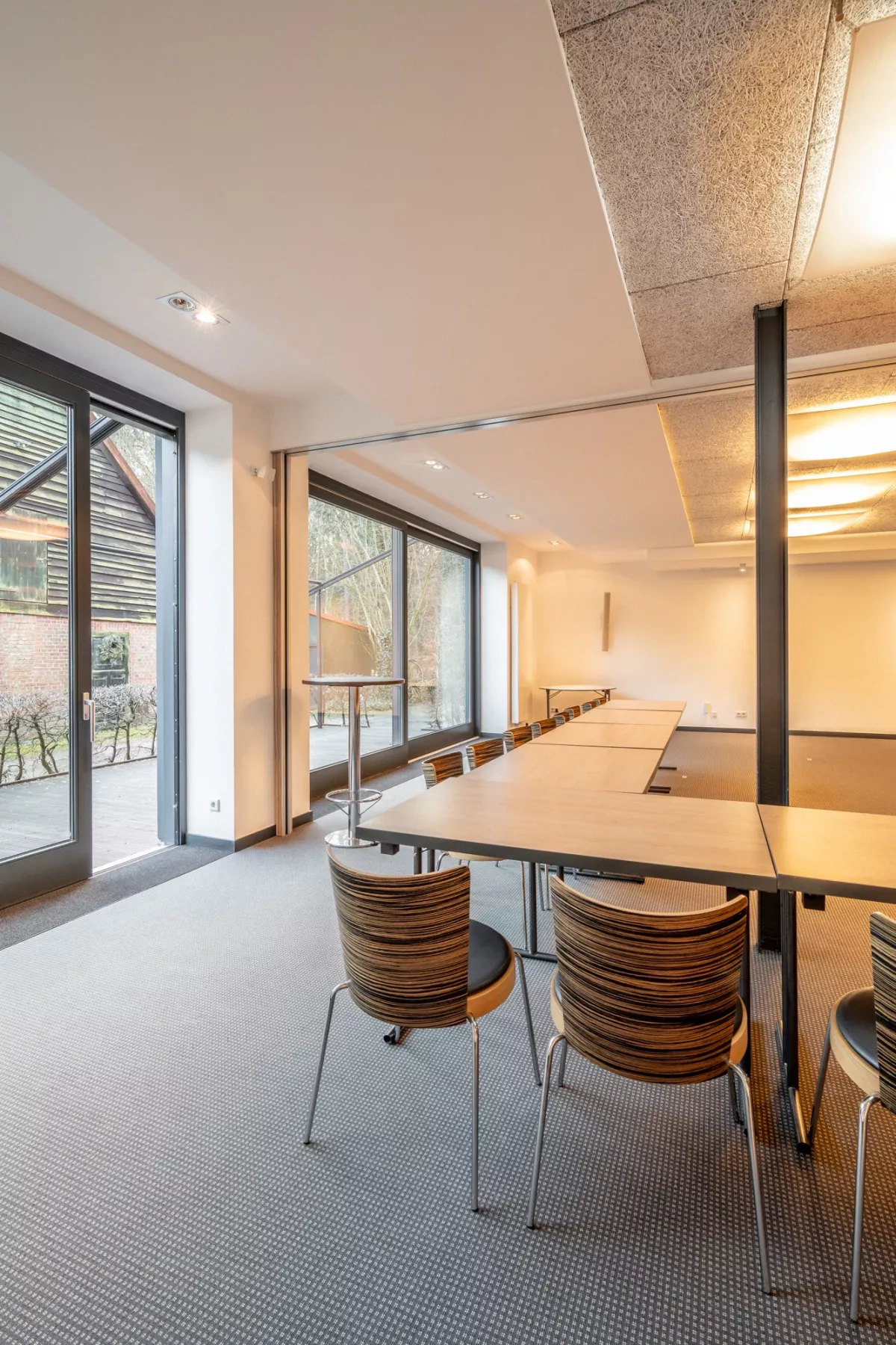 Moderner Veranstaltungsraum mit Holztisch, ergonomischen St&uuml;hlen und gro&szlig;en Glast&uuml;ren zum Au&szlig;enbereich, helles Licht von oben.