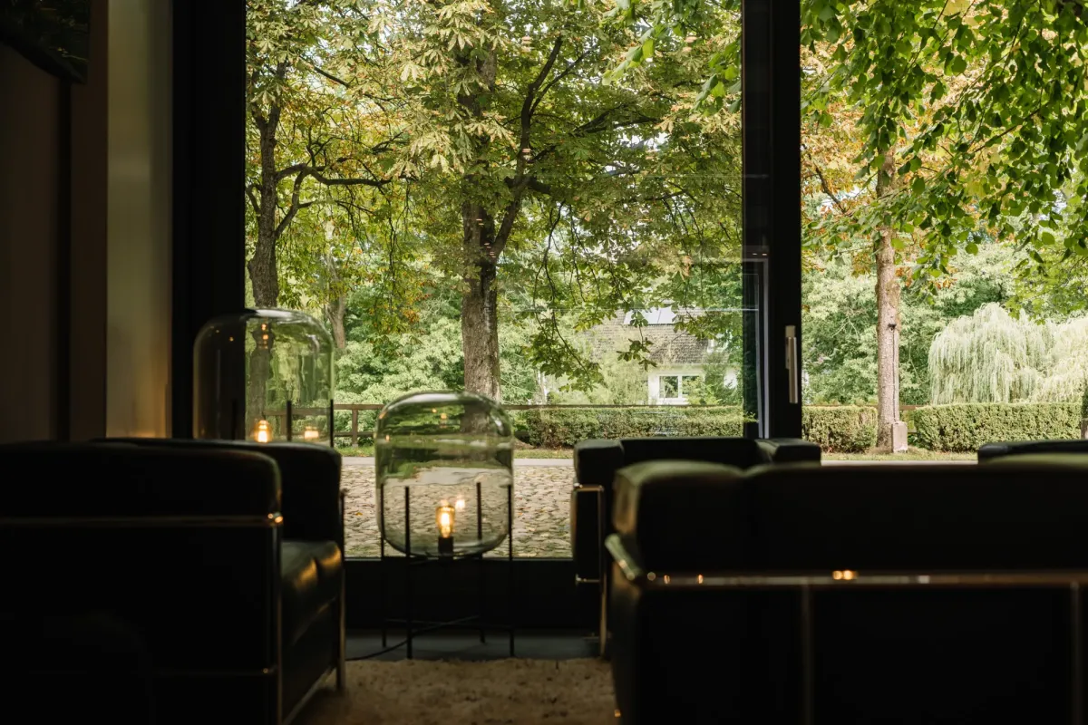 Moderne Lounge mit gro&szlig;en Panoramafenstern blickt auf Vorplatz und B&auml;ume, schwarze Ledersofas und moderne Lampe im Vordergrund.