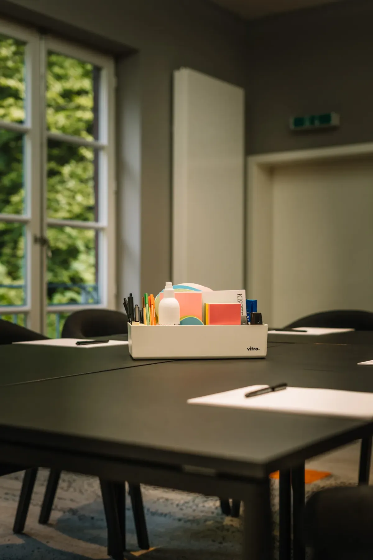Tisch in Seminarraum mit Stifthalter und B&uuml;romaterial im Tageslicht, Fenster mit Gr&uuml;nblick im Hintergrund.