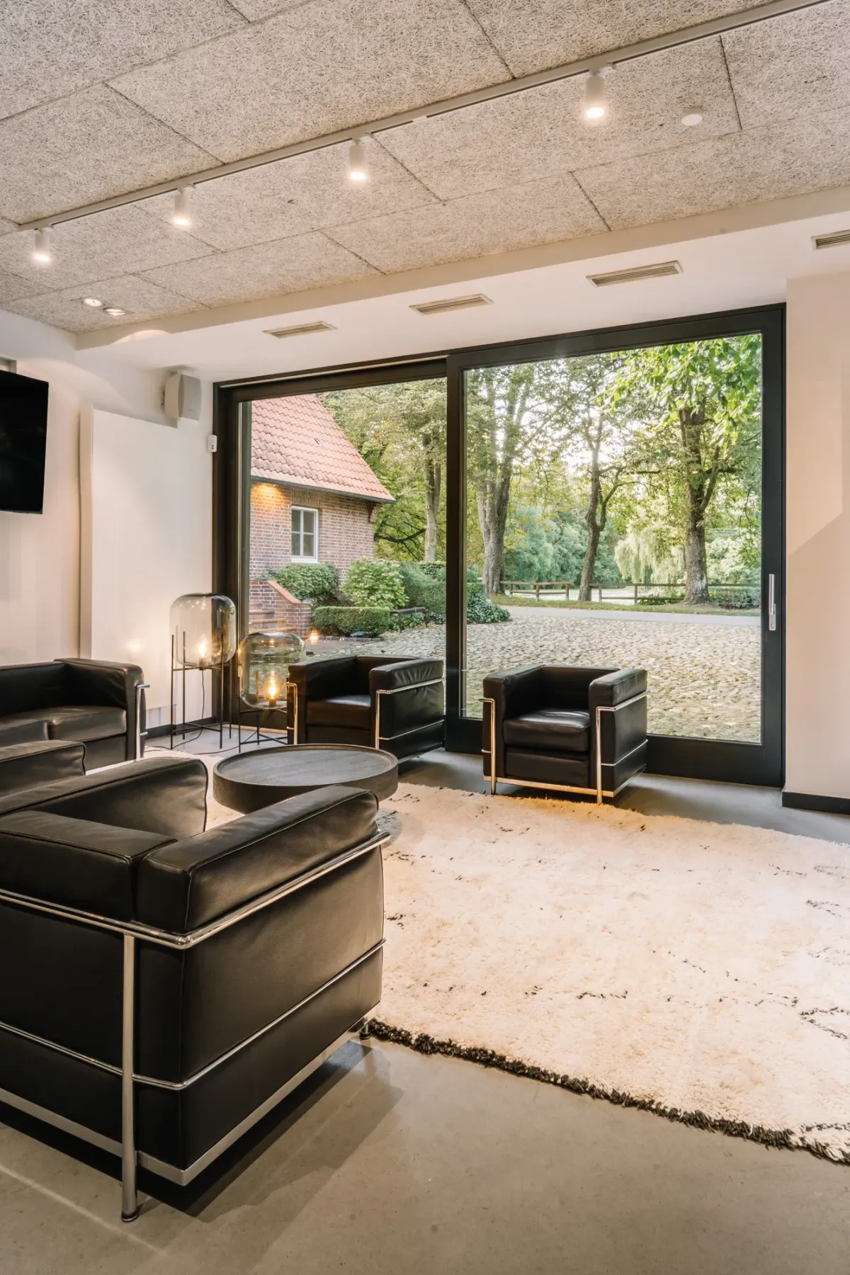 Moderner Lounge-Bereich mit schwarzen Ledersesseln, Betonboden und gro&szlig;en Glasschiebet&uuml;ren mit Blick auf Vorplatz und historisches Geb&auml;ude.