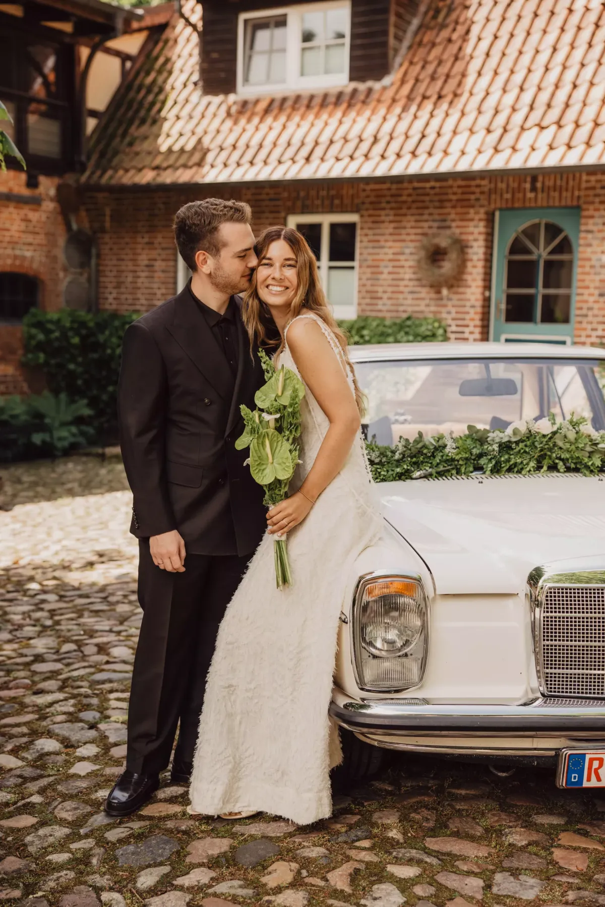 Brautpaar posiert vor einem vintage Hochzeitsauto vor einem Backsteinhaus. Der Br&auml;utigam tr&auml;gt einen schwarzen Anzug, die Braut ein wei&szlig;es Hochzeitskleid und h&auml;lt einen gr&uuml;nen Blumenstrau&szlig;.