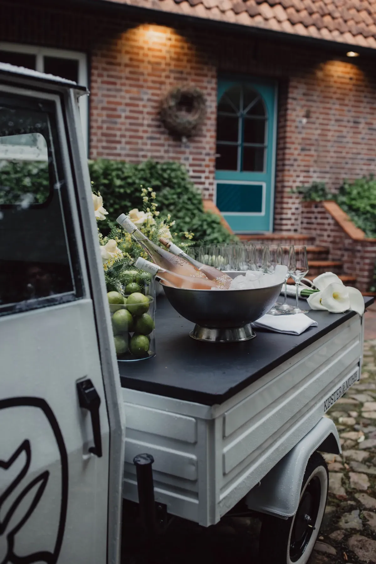 Ein mobile Sekttheke auf einem Anh&auml;nger vor einem Backsteinhaus mit t&uuml;rkisgr&uuml;ner T&uuml;r, bereit f&uuml;r Catering und Veranstaltungen.