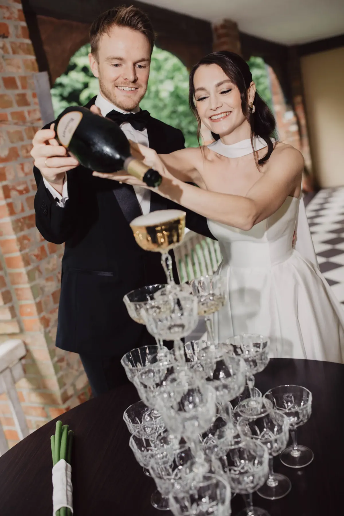 Brautpaar prostet mit Champagner zu, w&auml;hrend sie Champagner in Gl&auml;ser auf einem Tisch gie&szlig;en. Der Br&auml;utigam tr&auml;gt einen schwarzen Smoking, die Braut ein wei&szlig;es Kleid.