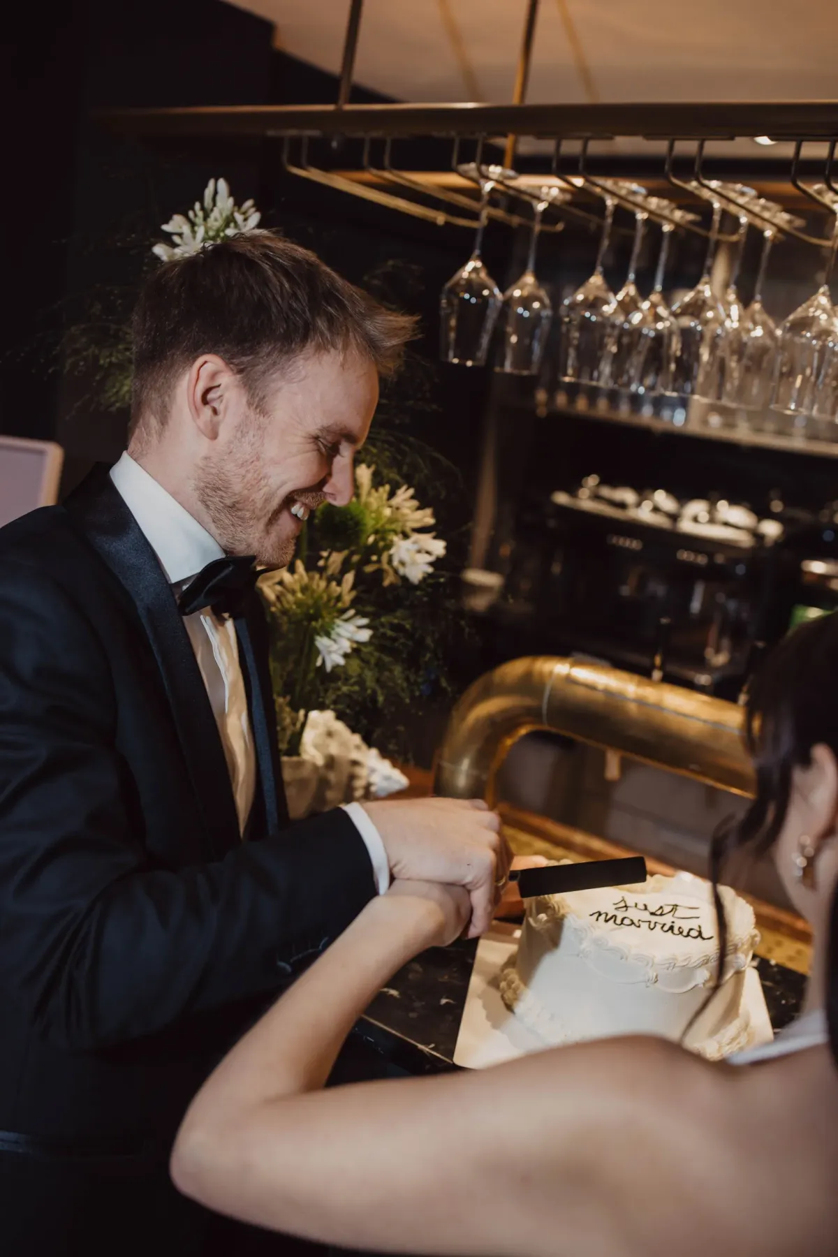 Br&auml;utigam in dunklem Anzug mit Fliege h&auml;lt gemeinsam mit Braut in wei&szlig;em Kleid ein Messer in der Hand und schneidet Hochzeitstorte an, im Hintergrund elegante Bar mit Gl&auml;sern.