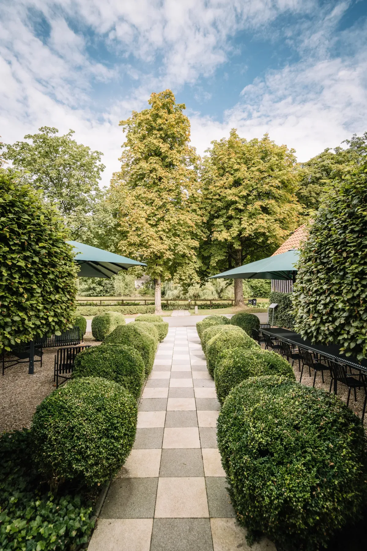 Formale Gartenanlage mit symmetrischen Buchsbaumhecken entlang eines Gehwegs, gro&szlig;en B&auml;umen und gro&szlig;e Schirme im Hintergrund unter blauem Himmel.