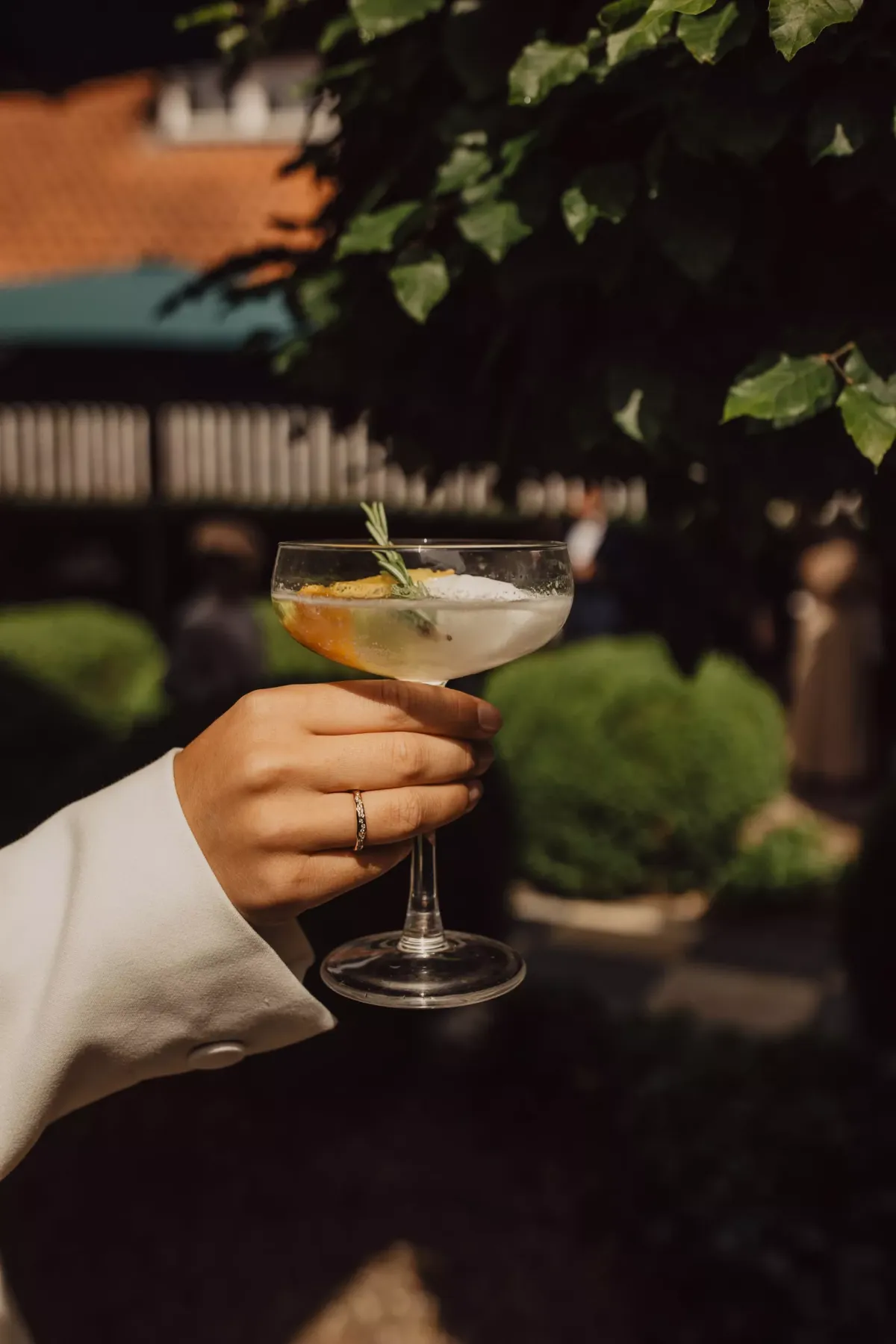 Hand in wei&szlig;em Anzug h&auml;lt Cocktailglas mit bernsteinfarbener Fl&uuml;ssigkeit und Eis vor gr&uuml;ner Gartenkulisse.