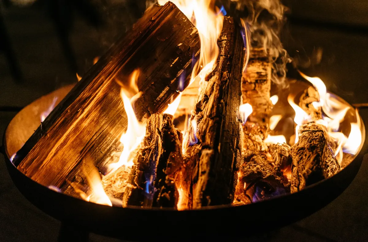 Loderndes Lagerfeuer mit Flammen in orangegoldenem Licht, umgeben von Dunkelheit und gl&uuml;henden Holzscheiten.