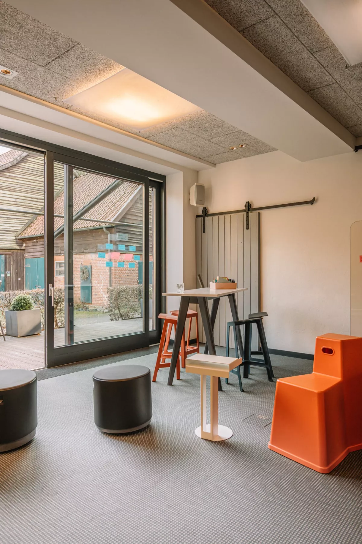 Moderner Tagungs- und Workshopraum mit gro&szlig;em Fenster zur Terrasse, Stehtischen, bunten Hockern und Sitzm&ouml;beln.