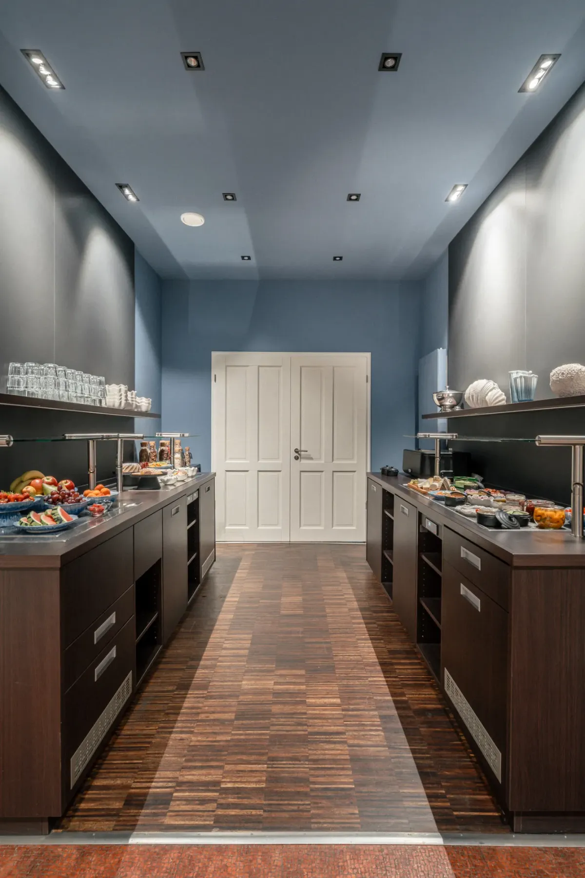 Moderne Buffetzone mit blauen W&auml;nden, dunklen Holzarbeitsplatten, Ausstellungstheken mit Speisen und wei&szlig;er T&uuml;r im Hintergrund.