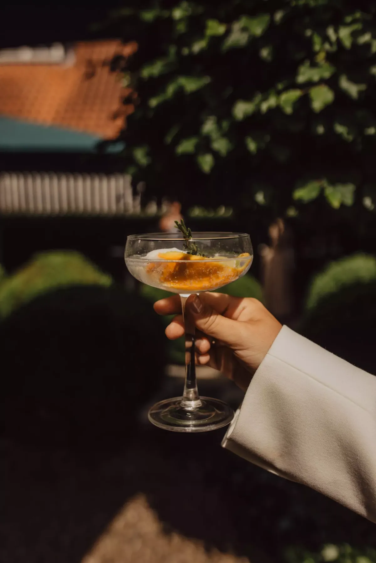 Hand in wei&szlig;em Anzug h&auml;lt Cocktailglas mit bernsteinfarbener Fl&uuml;ssigkeit und Eis vor gr&uuml;ner Gartenkulisse.