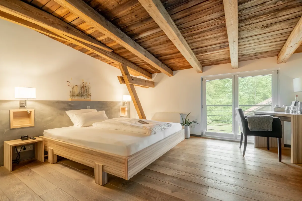 Helles Dachzimmer mit Holzbalkendecke, Doppelbett aus Holz, Nachttischen mit Lampen und gro&szlig;en Fenstern mit Ausblick ins Gr&uuml;ne.