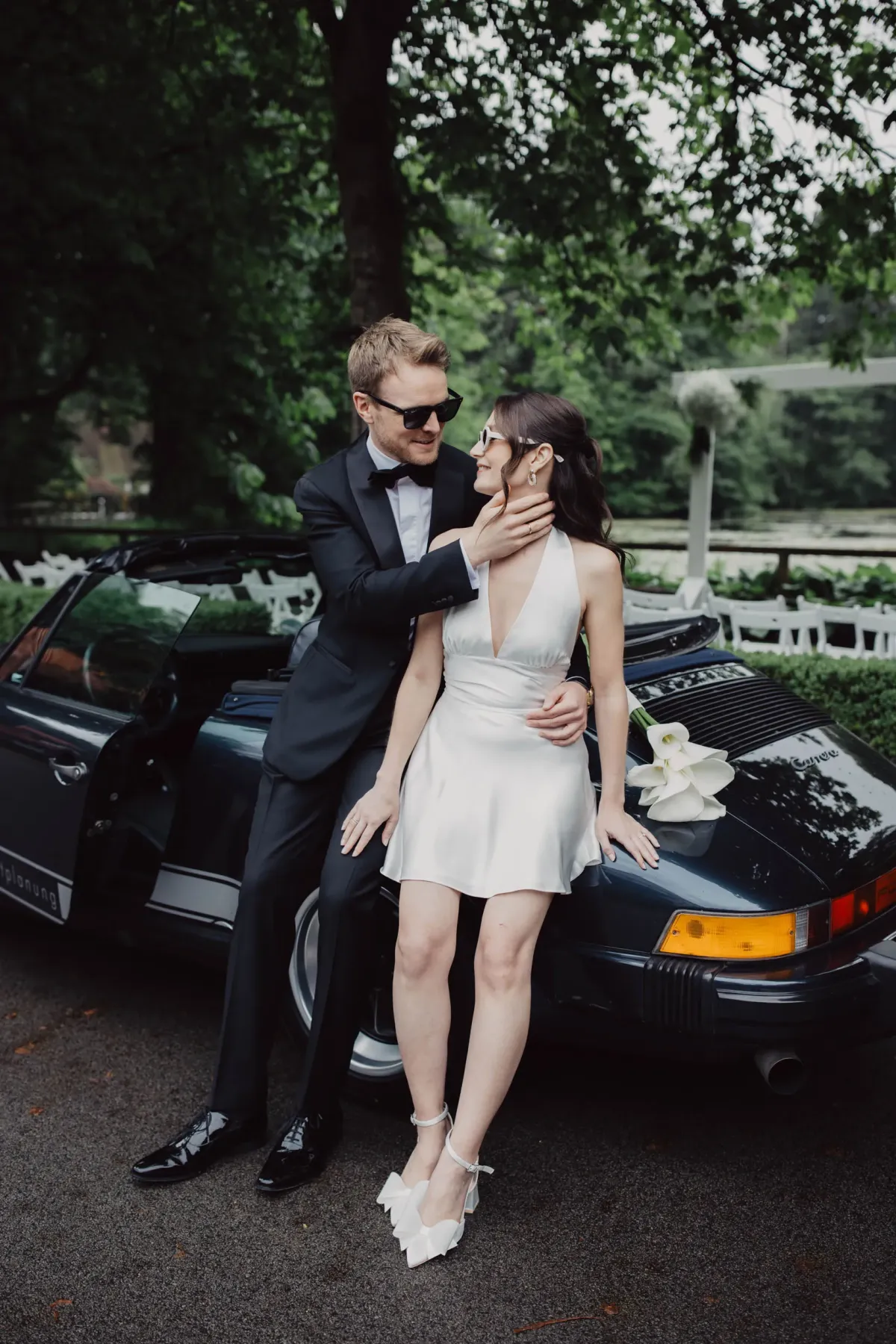 Elegantes Brautpaar steht vor einem schwarzen Oldtimer-Porsche an einer park&auml;hnlichen Location mit gr&uuml;nen B&auml;umen im Hintergrund.