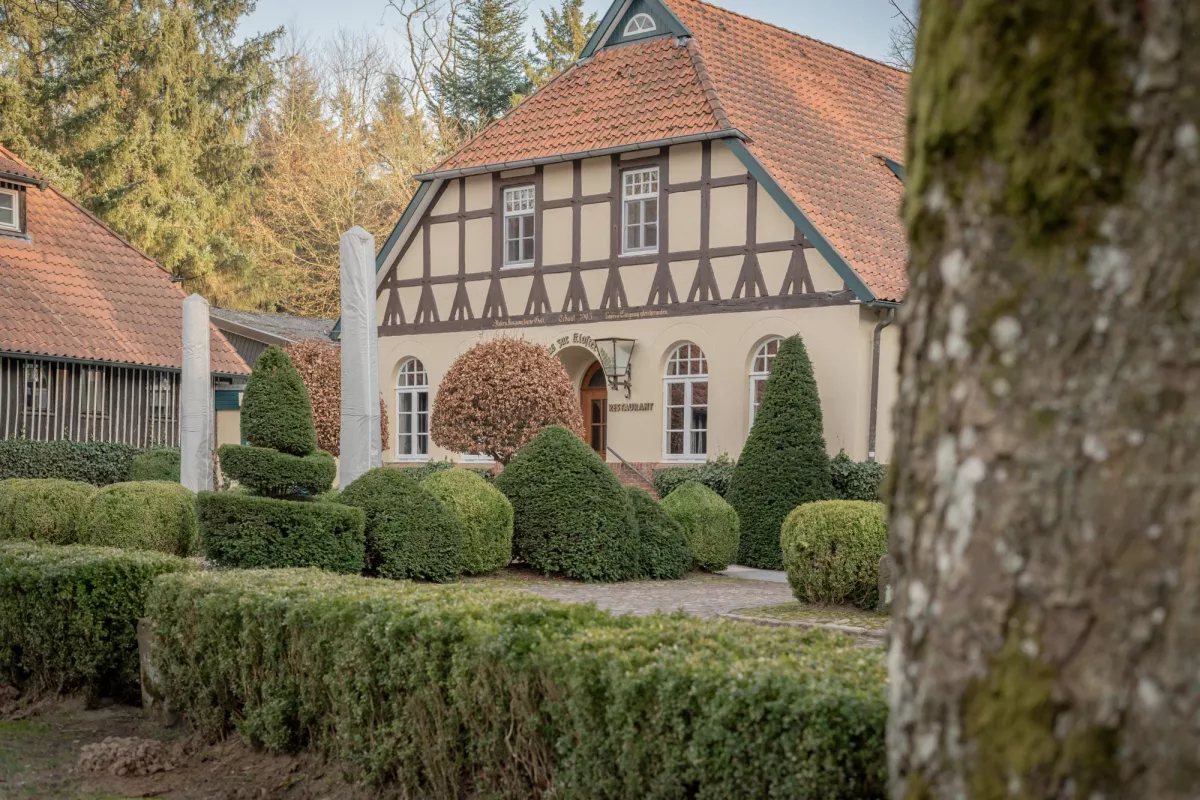 Fachwerkhaus mit Satteldach und roten Ziegeln, umgeben von geschnittenen Buchsb&auml;umen und Koniferen.