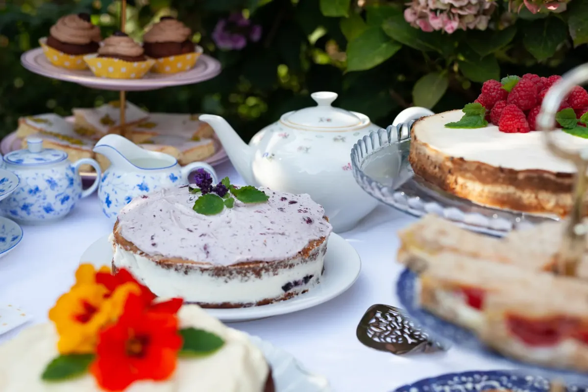 Elegante Kaffeetafel im Garten mit mehrschichtigen Torten, Geb&auml;ck, Blumendekorationen und Teeservice auf wei&szlig;em Tischtuch.