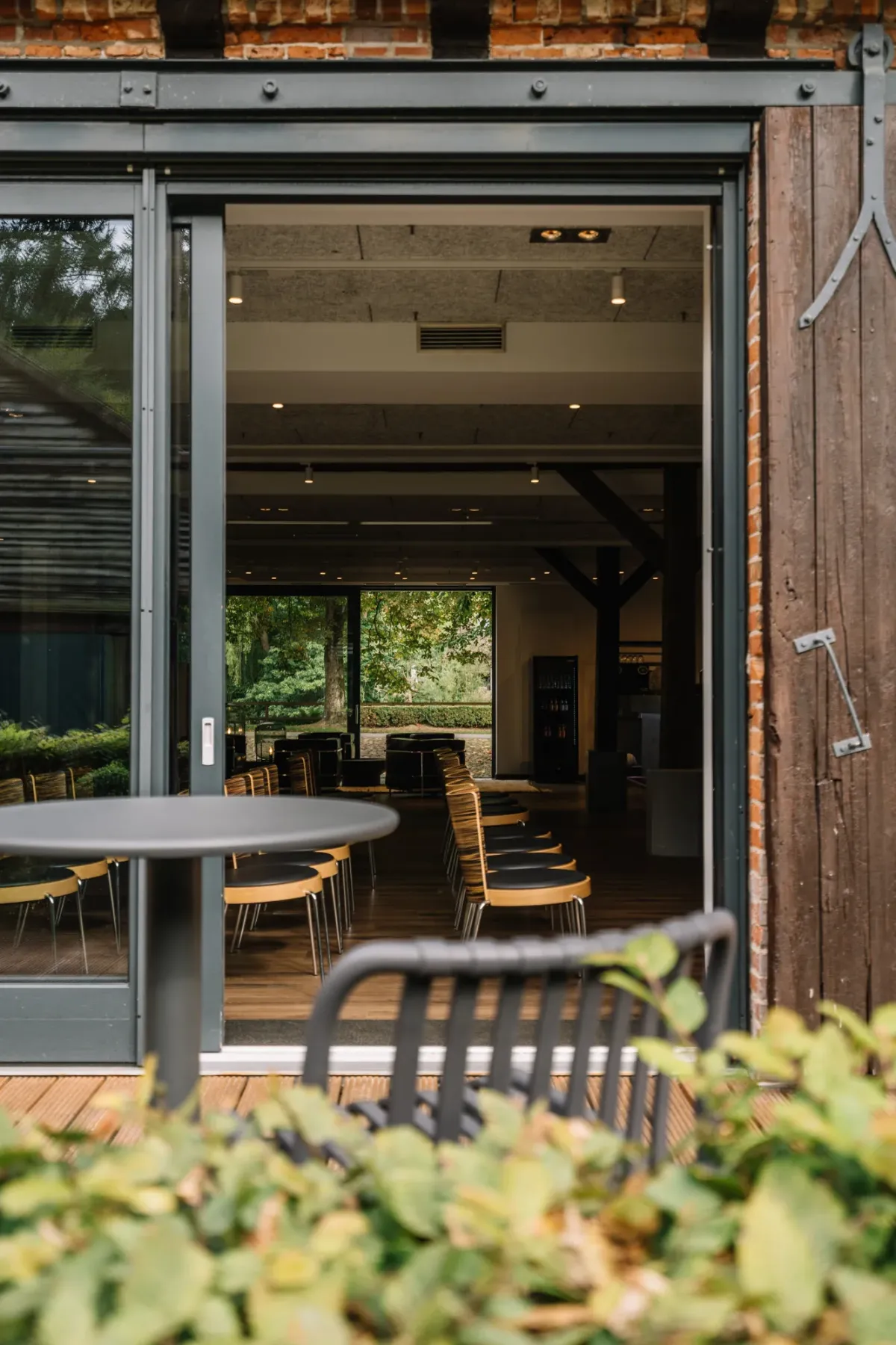 Moderne Schiebet&uuml;ren &ouml;ffnen den Blick auf einen eleganten Seminarraum mit Stuhlreihen und Durchgang ins Gr&uuml;ne im Hintergrund, Au&szlig;enterrasse mit gr&uuml;ner Hecke im Vordergrund.
