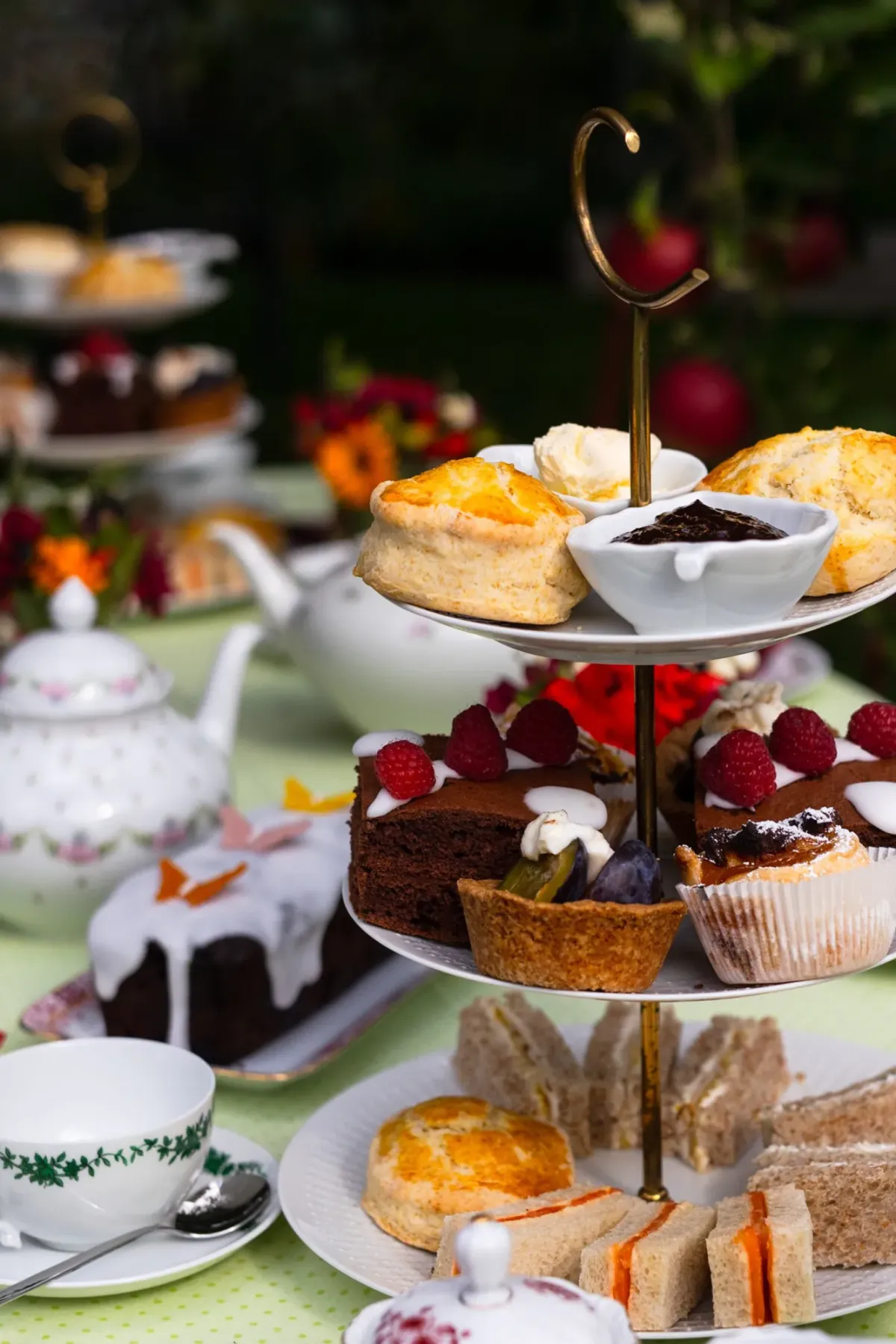 Mehrst&ouml;ckige Etagere mit Geb&auml;ck, Scones und Kuchen auf gr&uuml;nem Gartentisch, dekoriert mit roten Bl&uuml;ten f&uuml;r elegante Gartenfeier.