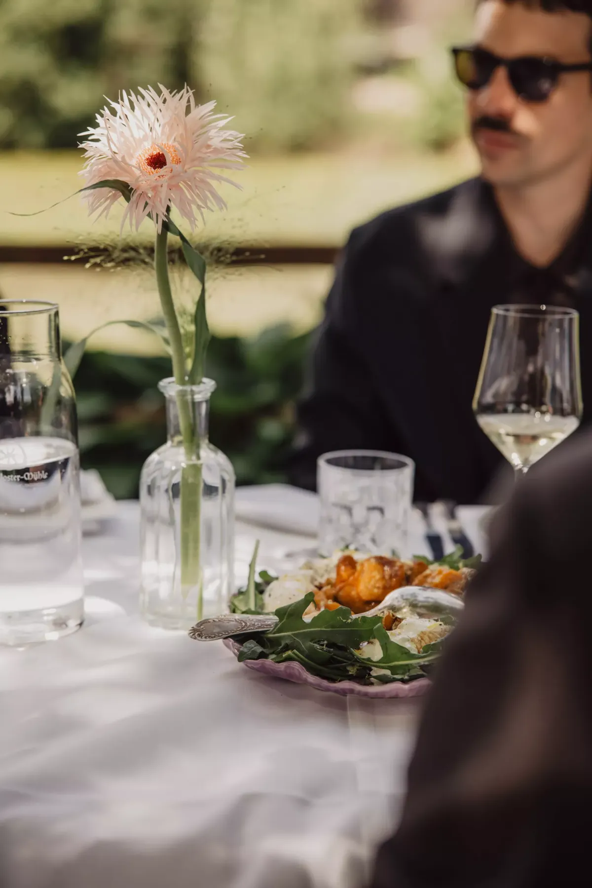 Ein elegantes Tischgedeck mit zartem rosa Gerbera in Glasvase, Weingl&auml;sern und Besteck, w&auml;hrend ein Mann im schwarzen Anzug und Sonnenbrille im unscharfen Hintergrund sitzt.