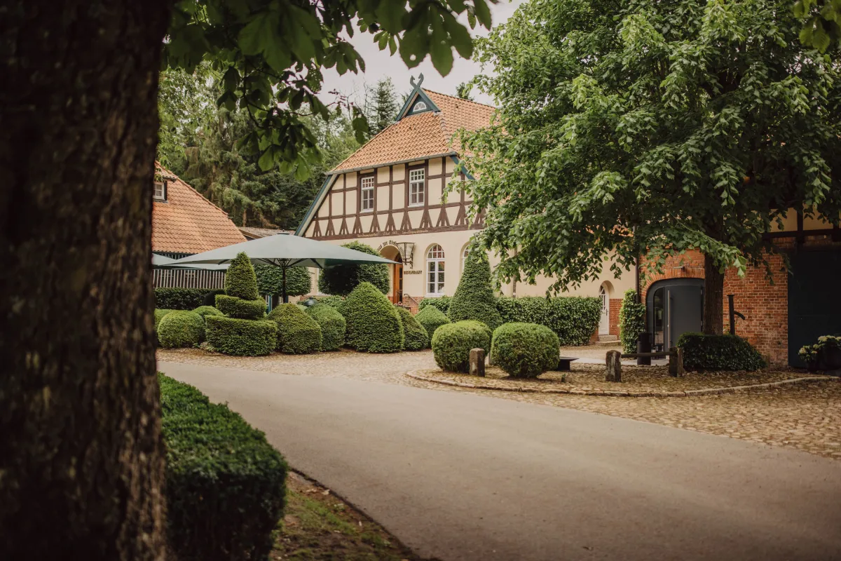 Historisches Fachwerkhaus mit Ziegeldach, umgeben von alten B&auml;umen und gepflegten Buchsb&auml;umen, mit wei&szlig;em Sonnensegel und Kiesweg im Vordergrund.