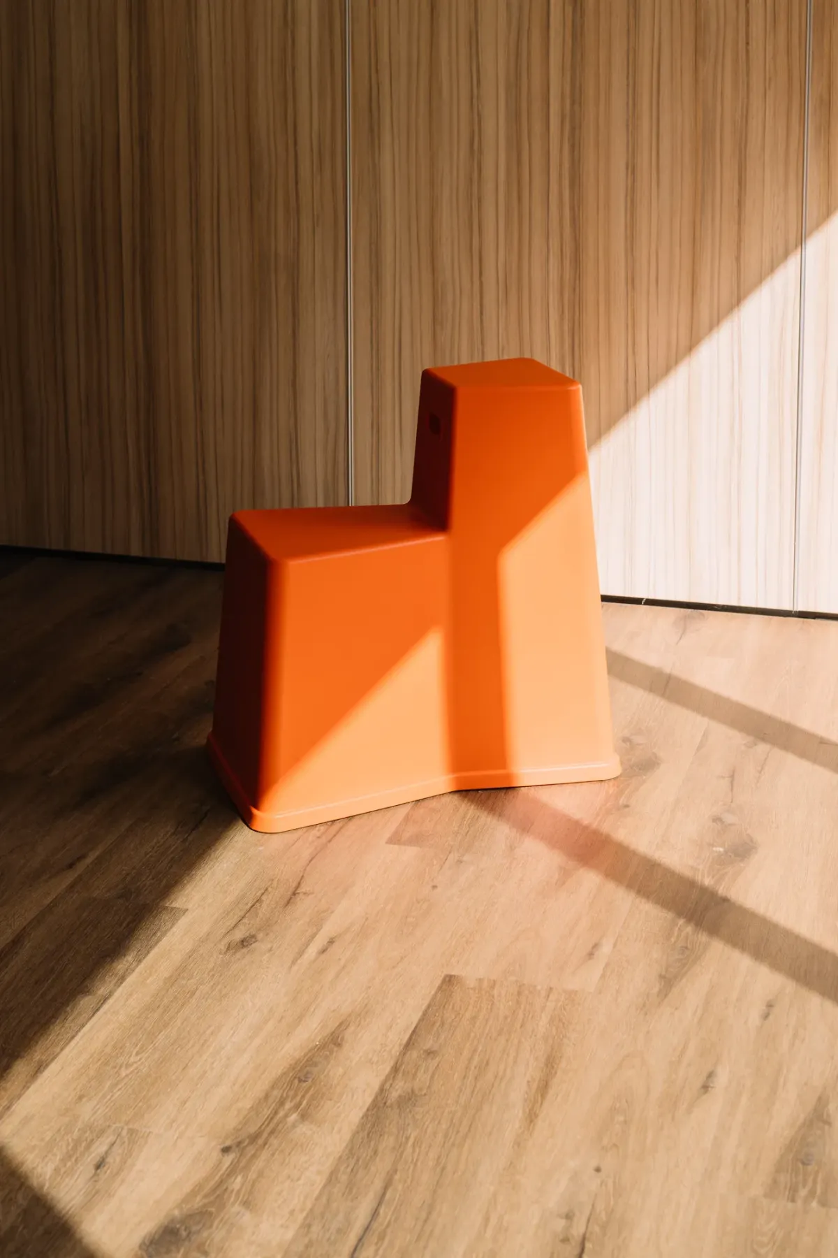 Orangefarbener moderner Stuhl mit geometrischer Form auf Holzdielen vor gestreifter Wandverkleidung.