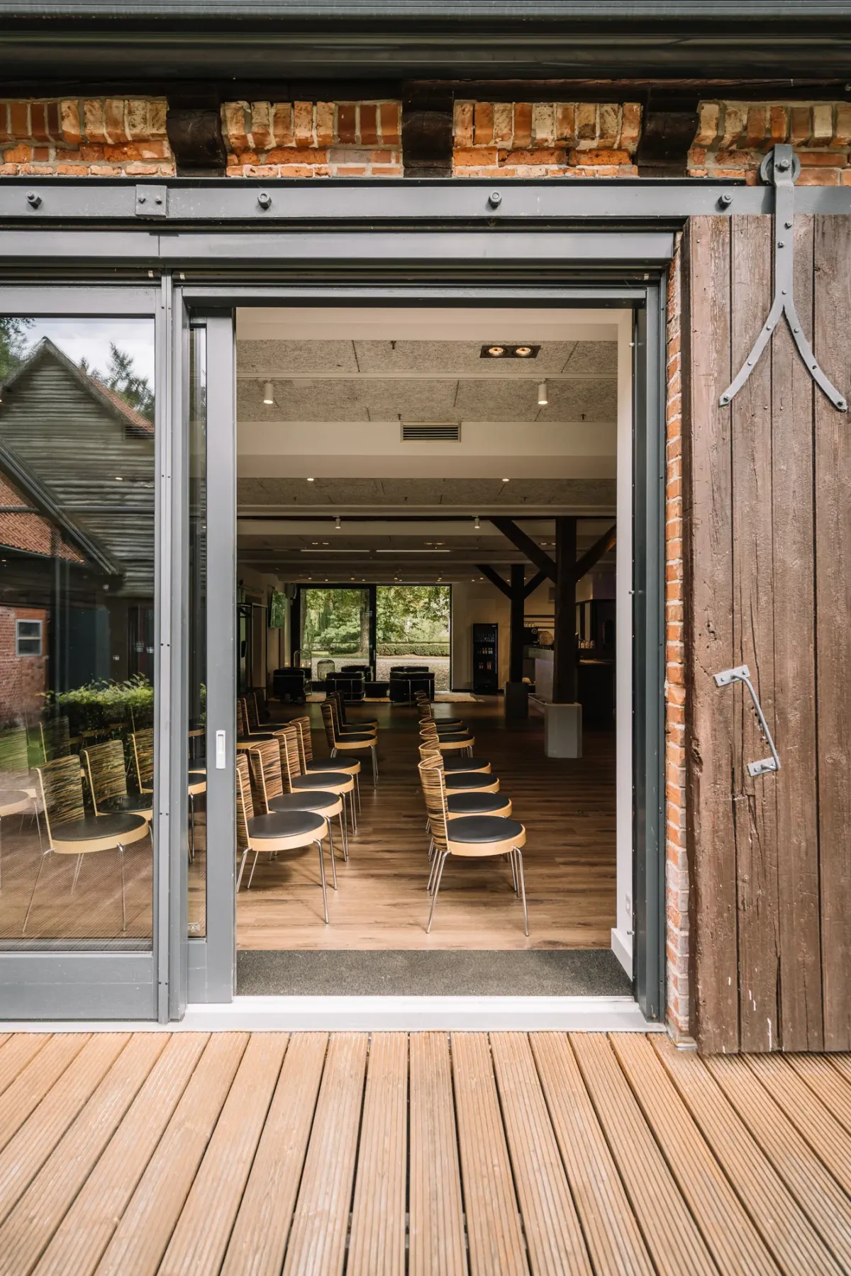 Blick durch gro&szlig;e Glasschiebet&uuml;ren in einen hellen Veranstaltungsraum mit Holzdielen, St&uuml;hlen und modernem Interieur. Au&szlig;en eine Holzterrasse mit heller Dielung. Naturnahes Ambiente f&uuml;r private Events und Feiern.