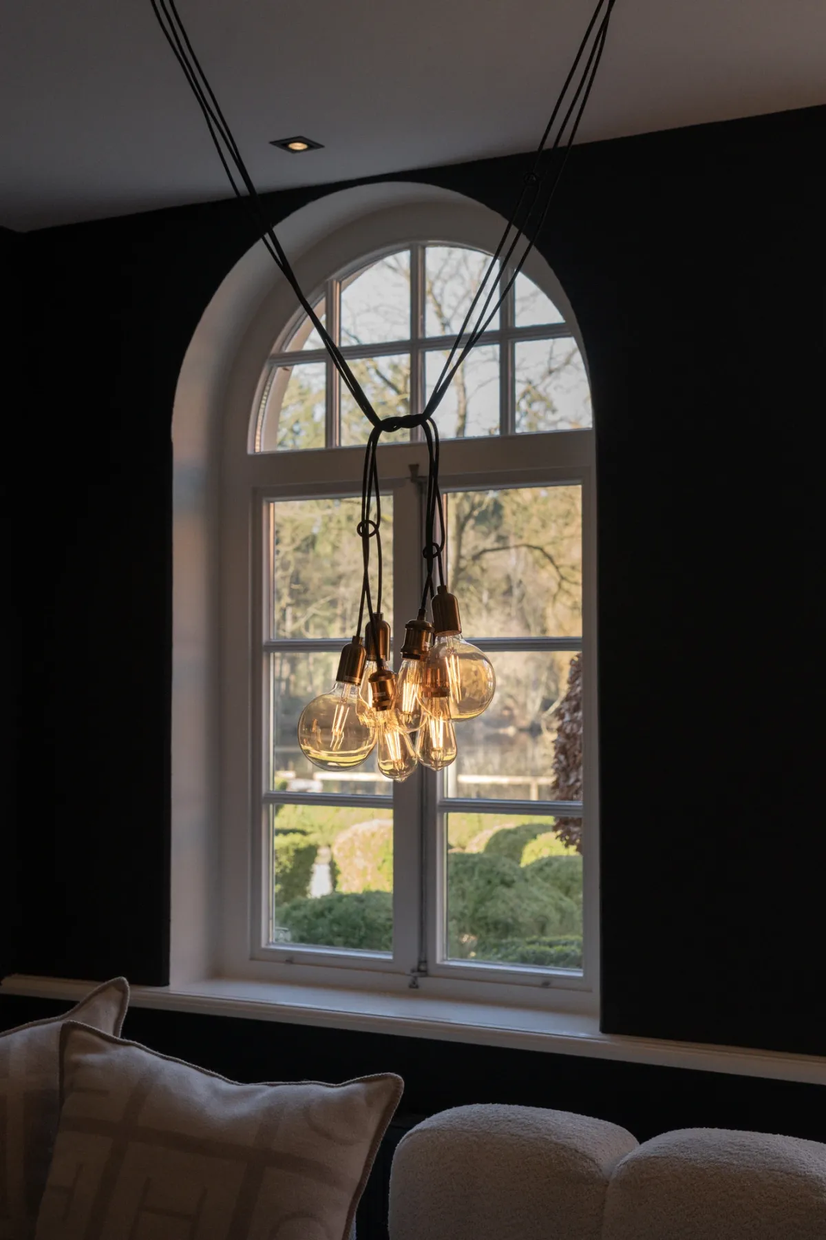 Gro&szlig;es Rundbogenfenster mit Ausblick auf gr&uuml;ne Landschaft, darunter h&auml;ngt eine moderne Pendelleuchte aus Vintage-Gl&uuml;hbirnen.
