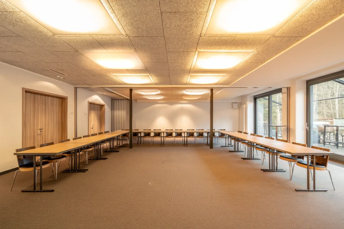 Gro&szlig;er heller Veranstaltungsraum mit Holzdecke, Reihen von Tischen und St&uuml;hlen, gro&szlig;en Fenstern und modernen Deckenleuchten.