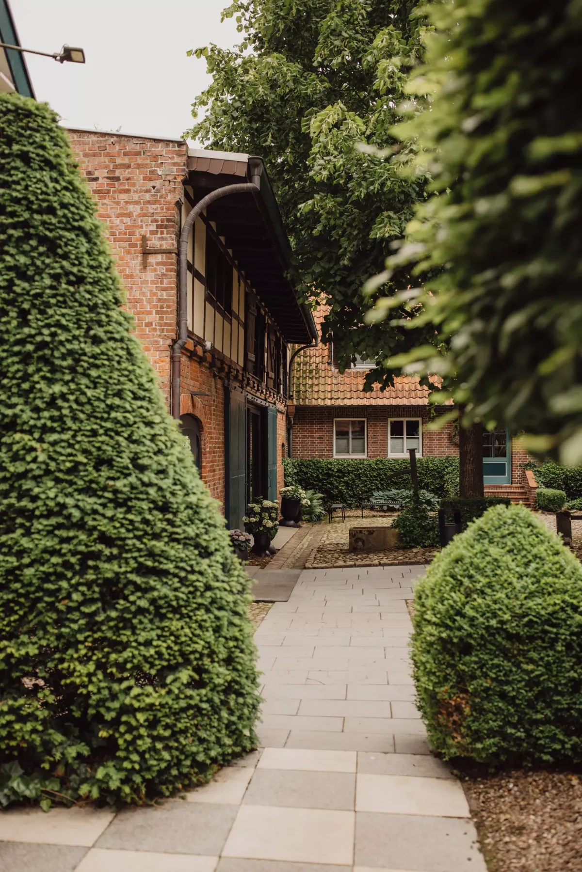 Gepflasterter Durchgang zwischen zwei Hecken f&uuml;hrt zu einem Backsteingeb&auml;ude mit Efeu bewachsener Fassade und Fachwerkdetails in einem gr&uuml;nen Innenhof.